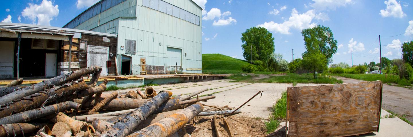 A pile of rusted black metal pipes on the ground with an abandoned corrugated metal building in the background.