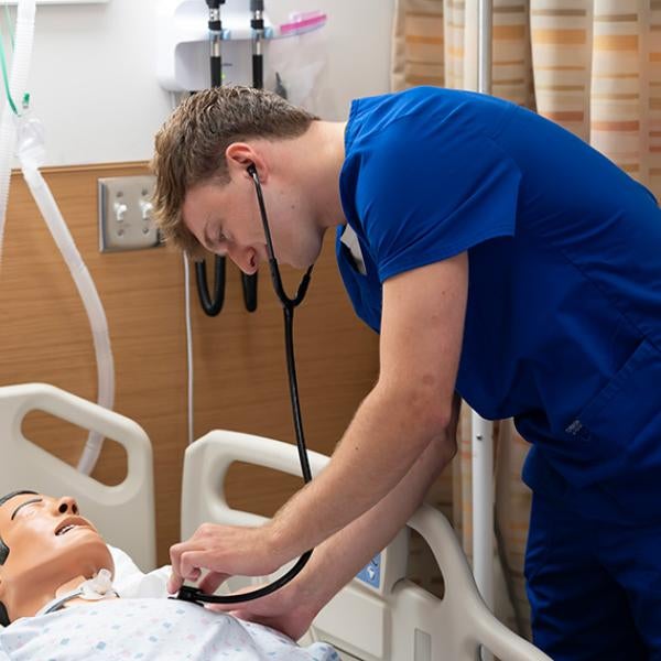 A student in scrubs holds a stethoscope to a dummy in a hospital bed