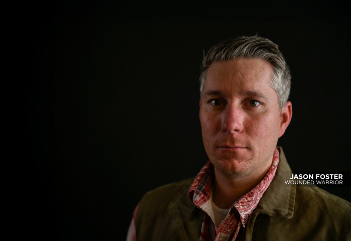 Wounded warrior Jason Foster is looking straight into the camera with serious expression. He is wearing a green vest over a red shirt. He is standing against a solid black background.