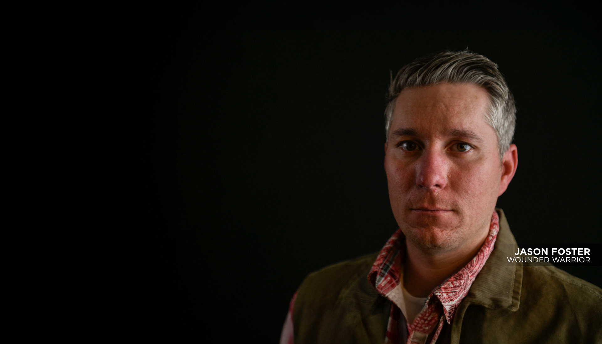 Wounded warrior Jason Foster is looking straight into the camera with serious expression. He is wearing a green vest over a red shirt. He is standing against a solid black background.