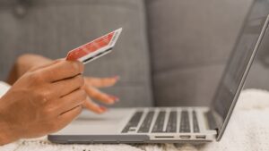 A photo of the hands of a person hovering over a laptop. One hand holds a credit card, suggesting the person is shopping online.