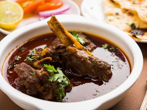 Bowl of lamb with garnishes and a curry- like consistency. There is naan in the background as well as a spoon next to the bowl