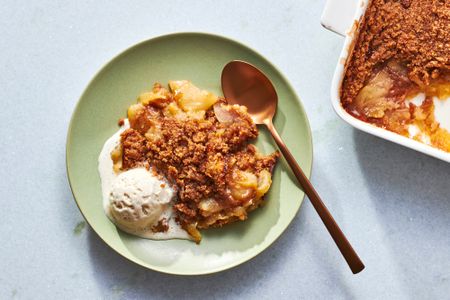 A serving of apple brown betty in a bowl, served with vanilla ice cream, with a baking dish of apple brown better on the side