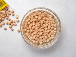 A large glass bowl with dried chickpeas soaking in water, with a bag of spilled dried chickpeas