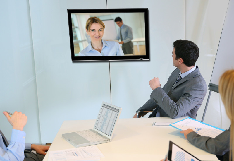 Drei Personen in einem Büro die auf einen Bildschirm mit weiteren Personen blicken und eine Videokonferenz abhalten. 