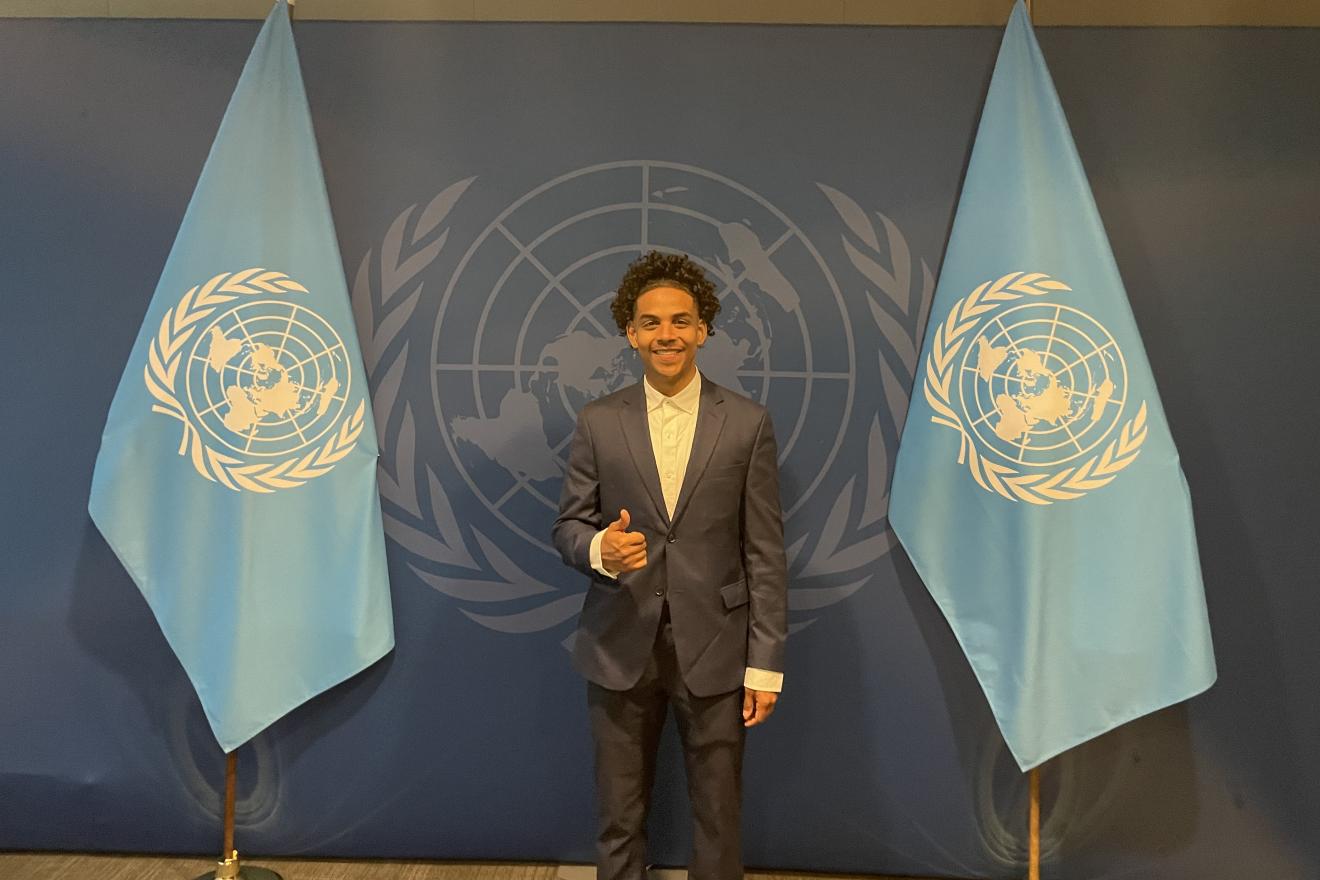 A young man stands in front of two UN flags.