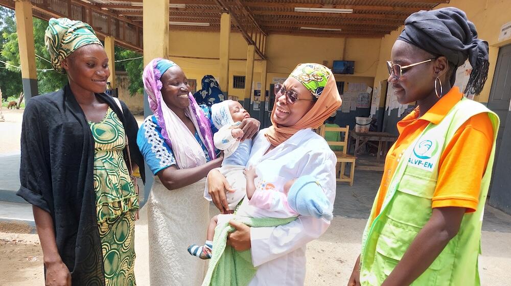 Four women stand laughing outside a building, one holds two newborn babies.