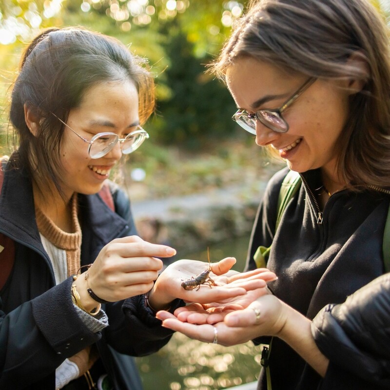 Two students standing together with one holding a small crayfish in their hands while they both are smiling