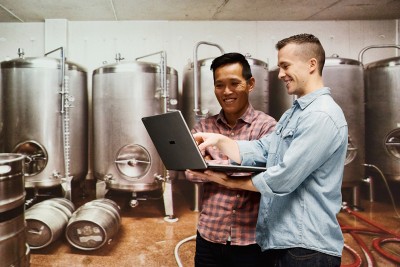 Two people are smiling in an industrial kitchen while looking and pointing at a computer screen.