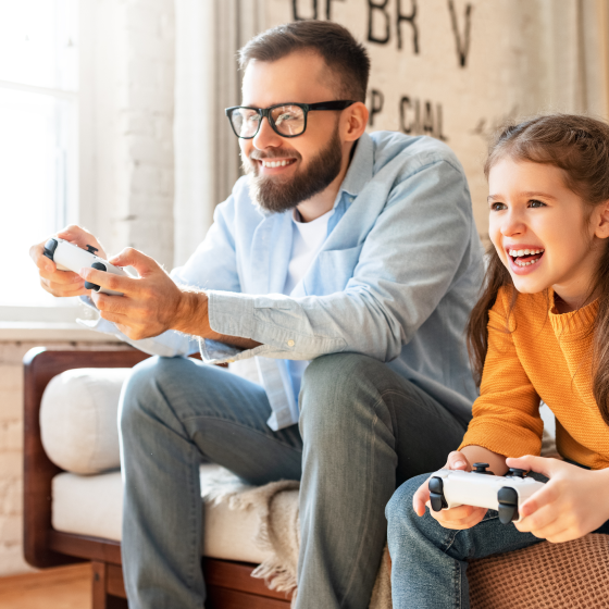 Man with a black beard and black rimmed glasses wearing a blue shirt with white t-shirt underneath and blue jeans sitting on a sofa alongside a young girl with long brown hair wearing an orange jumper and blue jeans while playing a computer game