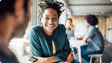 People in a coffee shop, the person in the front is sitting at a table at a coffee table and smiling.