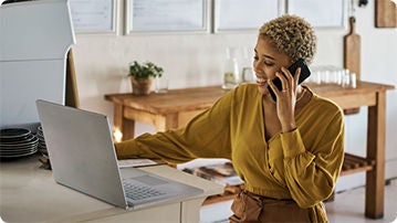 A person sitting indoors at a table with a laptop and talking on a phone.