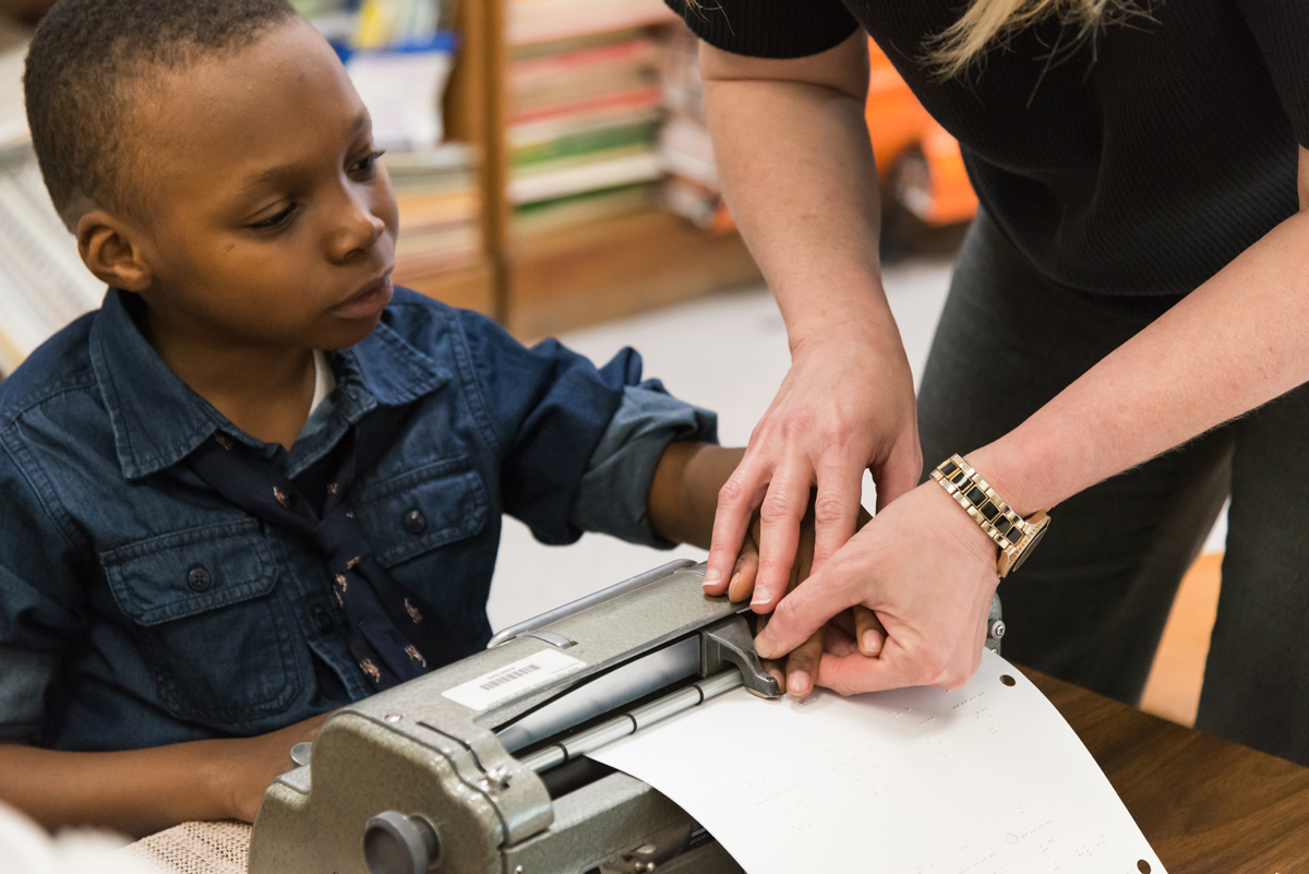 A young Black boy uses a braille typewriter, guided by his teacher.