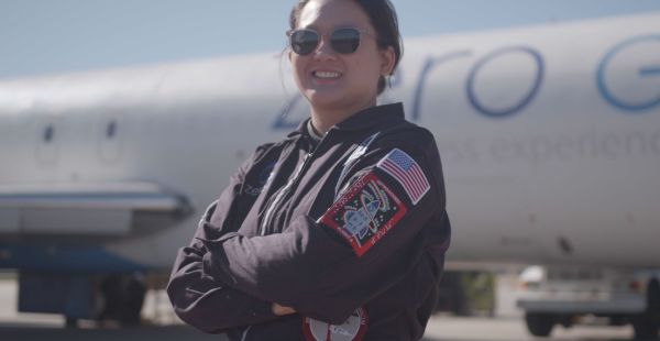 A woman in a flight suit and sunglasses stands with her arms crossed. Patches on one sleeve show an American flag and a rocket ship. A plane is visible in the background.