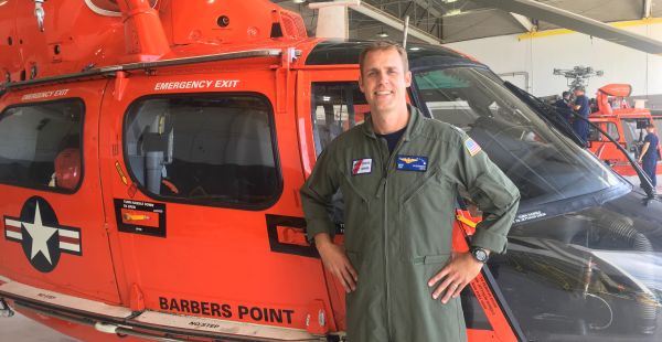 Gene McGuinness stands in front of a red helicopter wearing an olive flightsuit with a Coast Guard patch on it.