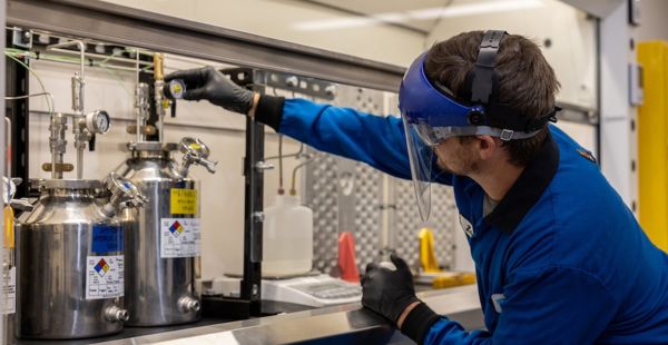 Zach Goodman, wearing full lab protection, adjusts a gauge above a stainless steel cylinder in a lab.