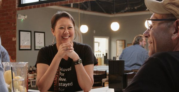 Jennifer Yang stands in her craft distillery's tasting room in Westminster, Maryland. She smiles with her elbows on the bar and her hands clasped. Behind Yang, you can see more of the restored historic venue. You can glimpse part of the company t-shirt Yang is pictured wearing. Vertically it reads, "Westminster." Horizontally it reads, "Moved for love, stayed for whiskey." 