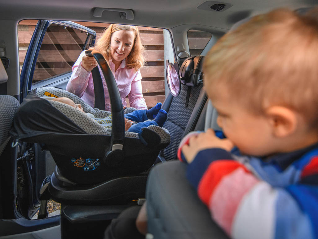 A woman putting an infant car seat in the car, while a toddler in a front-facing seat watches