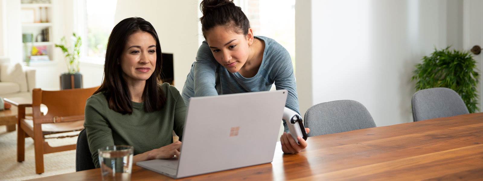 A mother using her a laptop alongside her daughter holding an Xbox controller