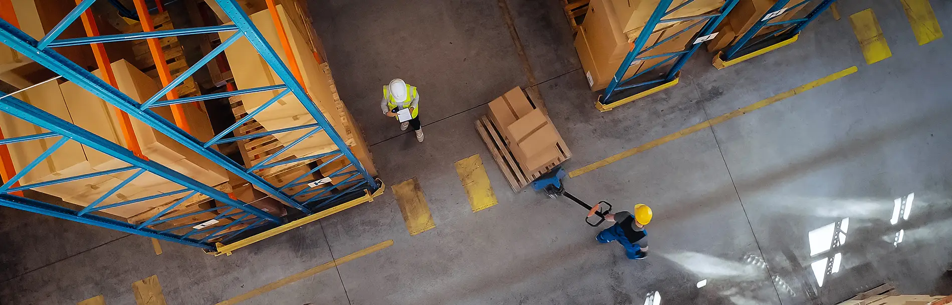 Two warehouse workers wearing PPE, moving boxes with pallet jack, and practicing proper health and safety protocols.