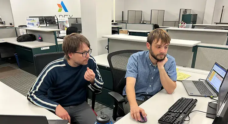 Two Apex Companies team members in an office having a discussion at a desk in front of a computer