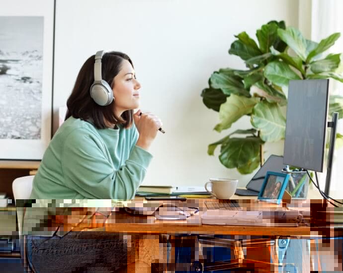 Woman wearing headphones, holding pen and looking at a PC monitor