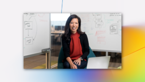 Portrait of female executive in red shirt, smiling and facing camera. She is in an office conference room setting, using an open laptop (screen not shown). Two whiteboards are partially visible in the background, with diagrams and flowcharts written on them.