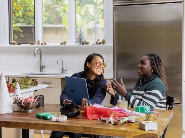 Two women in a kitchen, smiling as they refer to a laptop computer as they make holiday ornaments