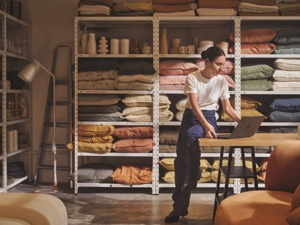 Woman leaning against a table working on a laptop next to shelves of textiles