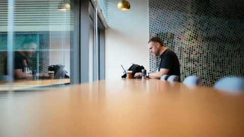 Tobias Brick, a man with a social disability, sits alone in the corner of an office building and works on a laptop. Accessibility, disability, and inclusion collection.