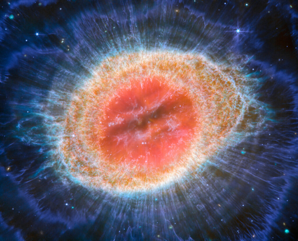  This image of the Ring Nebula appears as a distorted doughnut. The nebula’s inner cavity hosts shades of red and orange, while the detailed ring transitions through shades of yellow in the inner regions and blue/purple in the outer region. The ring’s inner region has distinct filament elements.