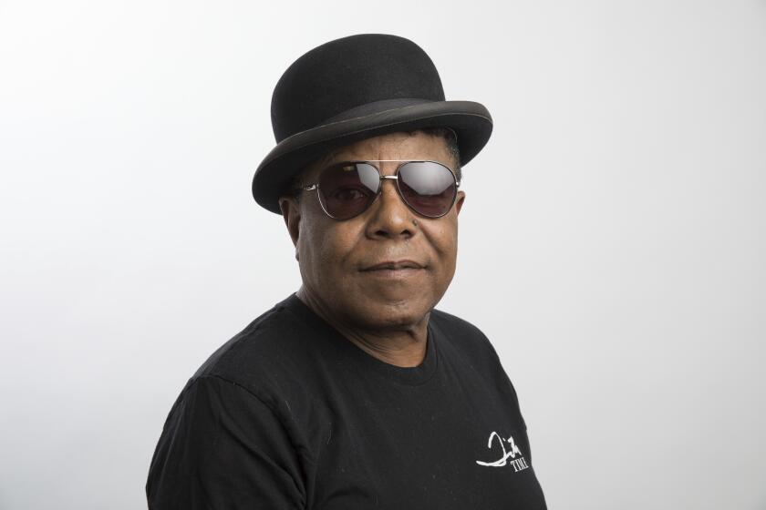 Tito Jackson poses for a portrait in a black t-shirt, dark aviator glasses and bowler hat