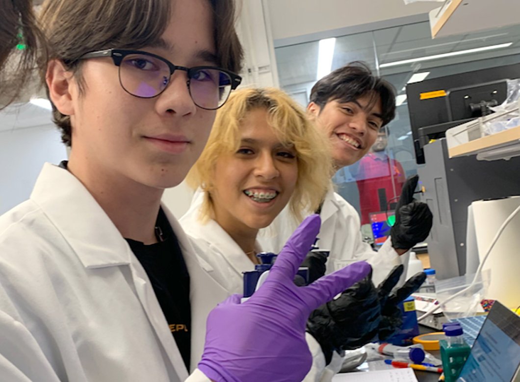 Four students are working in a scientific lab wearing rubber gloves, and using scientific instruments and a computer. All are smiling and facing the camera.