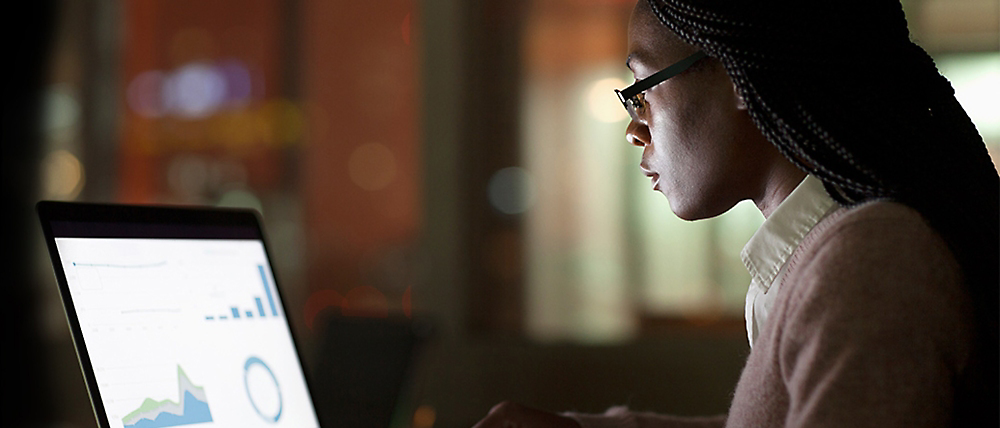 En kvinne med briller som fokuserer konsentrert på analyse av diagrammer på skjermen på en bærbar datamaskin, i et sparsommelig opplyst rom.