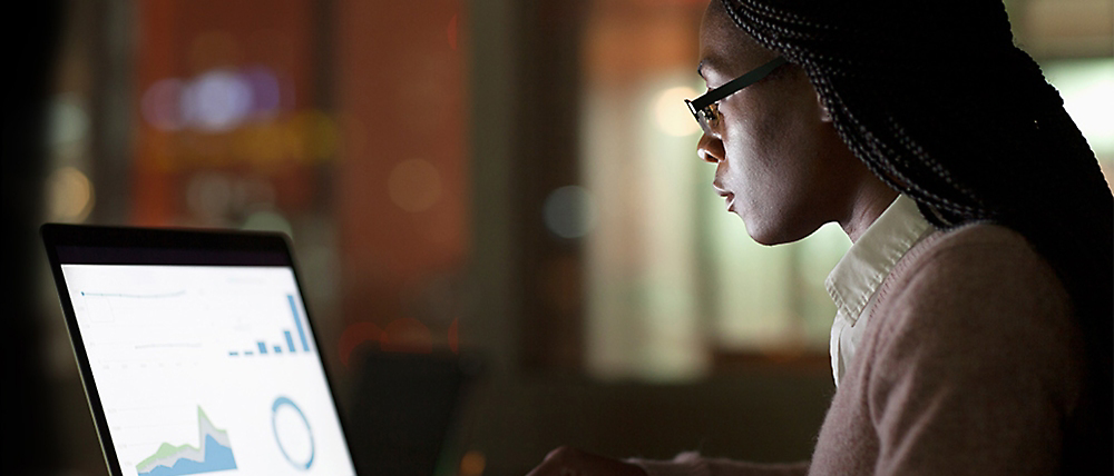 Een vrouw met een bril richt zich aandachtig op het analyseren van grafieken op haar laptopscherm in een zwak verlichte ruimte.