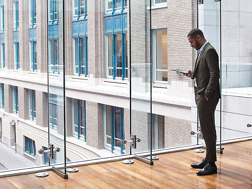 En forretningsmann i dress ser på en smarttelefon, ved siden av en glassvegg over en bygate med kontorbygg.