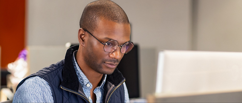 A man wearing glasses and a blue shirt with a dark vest is focused on his work in an office setting.