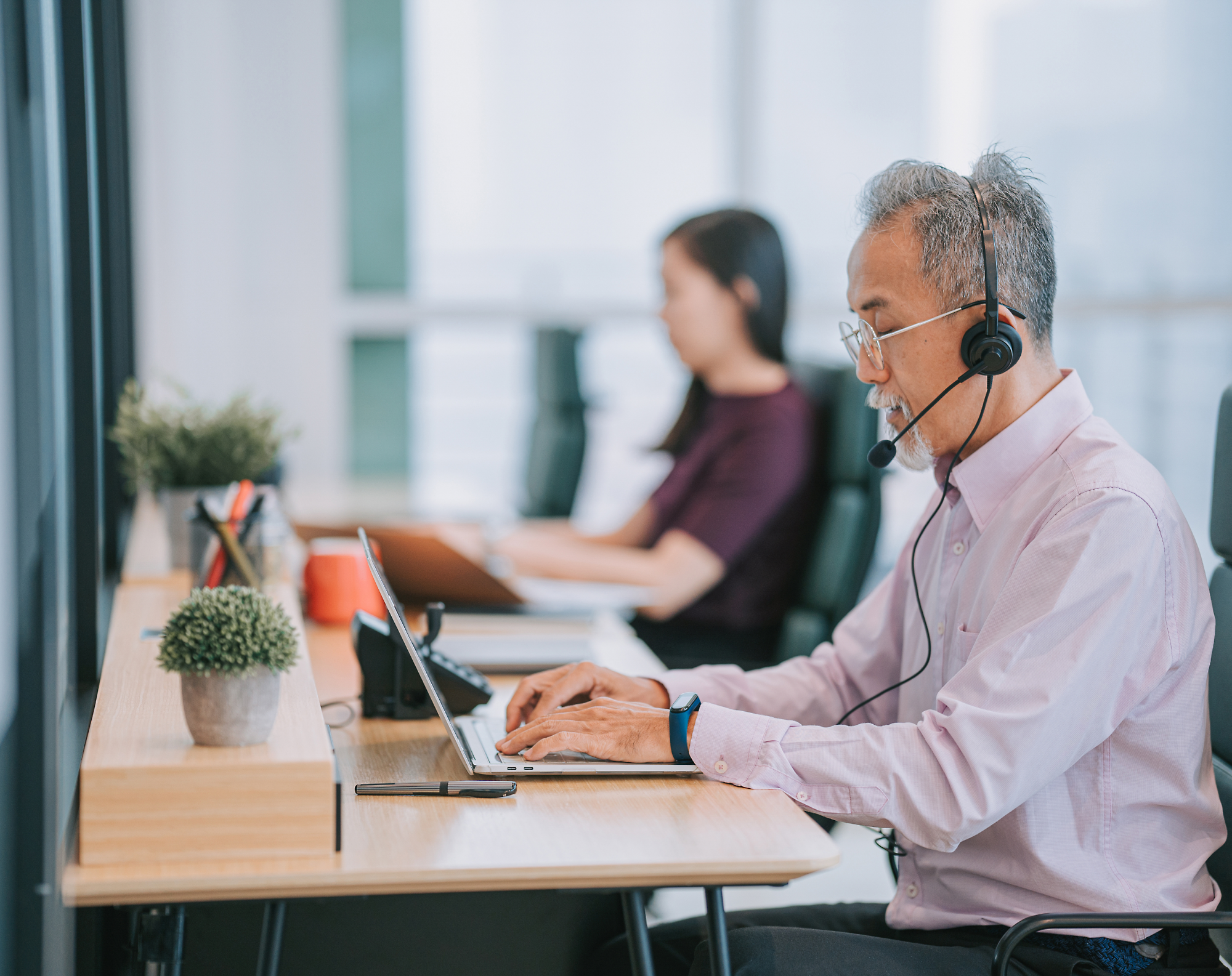 Two office workers sit at desks, wearing headsets, and working on laptops.