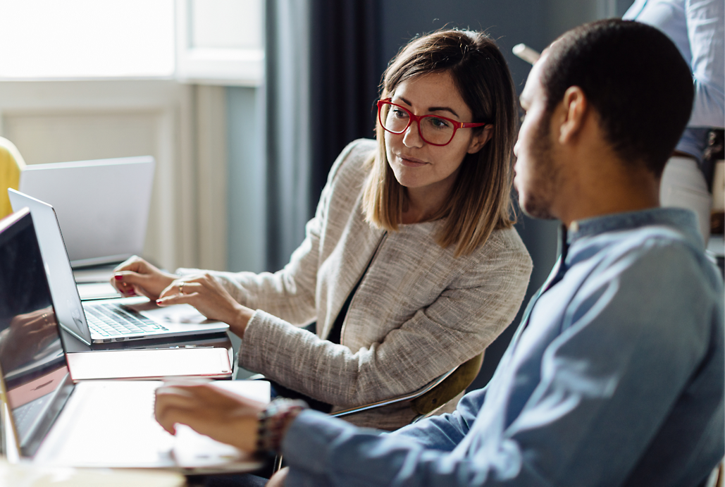 Two professionals, a woman with glasses and a man, using laptops and discussing work in an office setting.