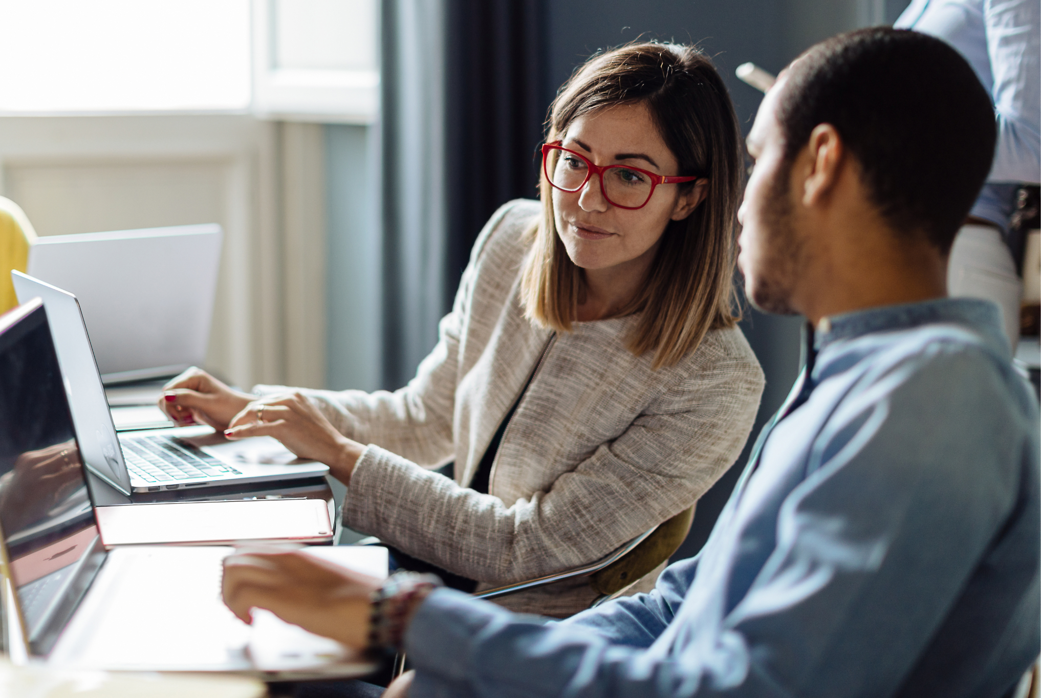 Two professionals, a woman with glasses and a man, using laptops and discussing work in an office setting.