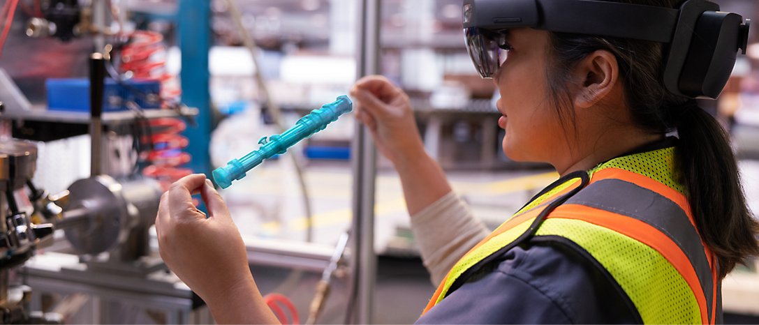 En medarbejder iført sikkerhedsvest og beskyttelsesbriller undersøger en blå mekanisk del i et industrielt miljø.