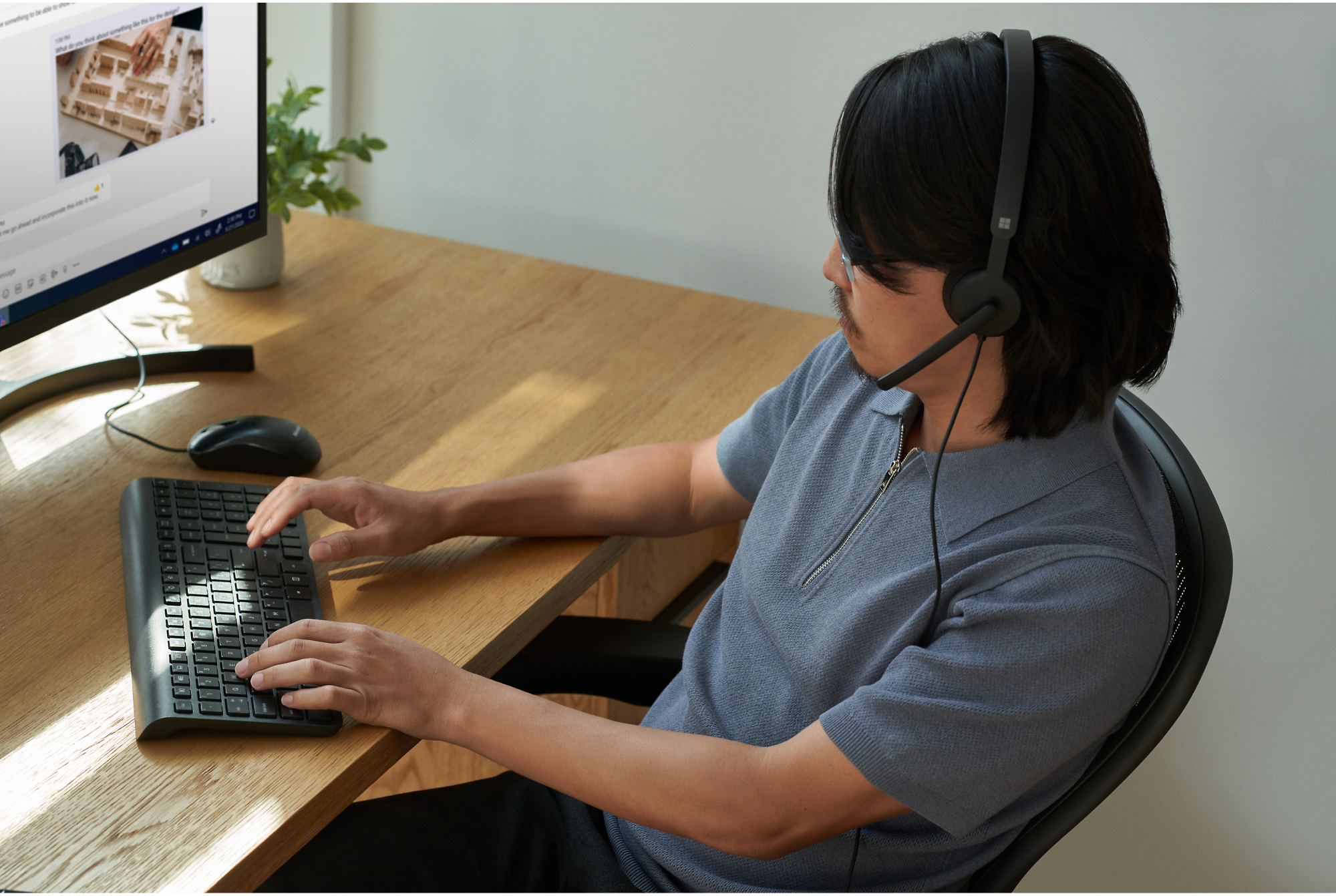 A person wearing a headset sits at a desk, typing on a keyboard, and looking at a computer monitor.