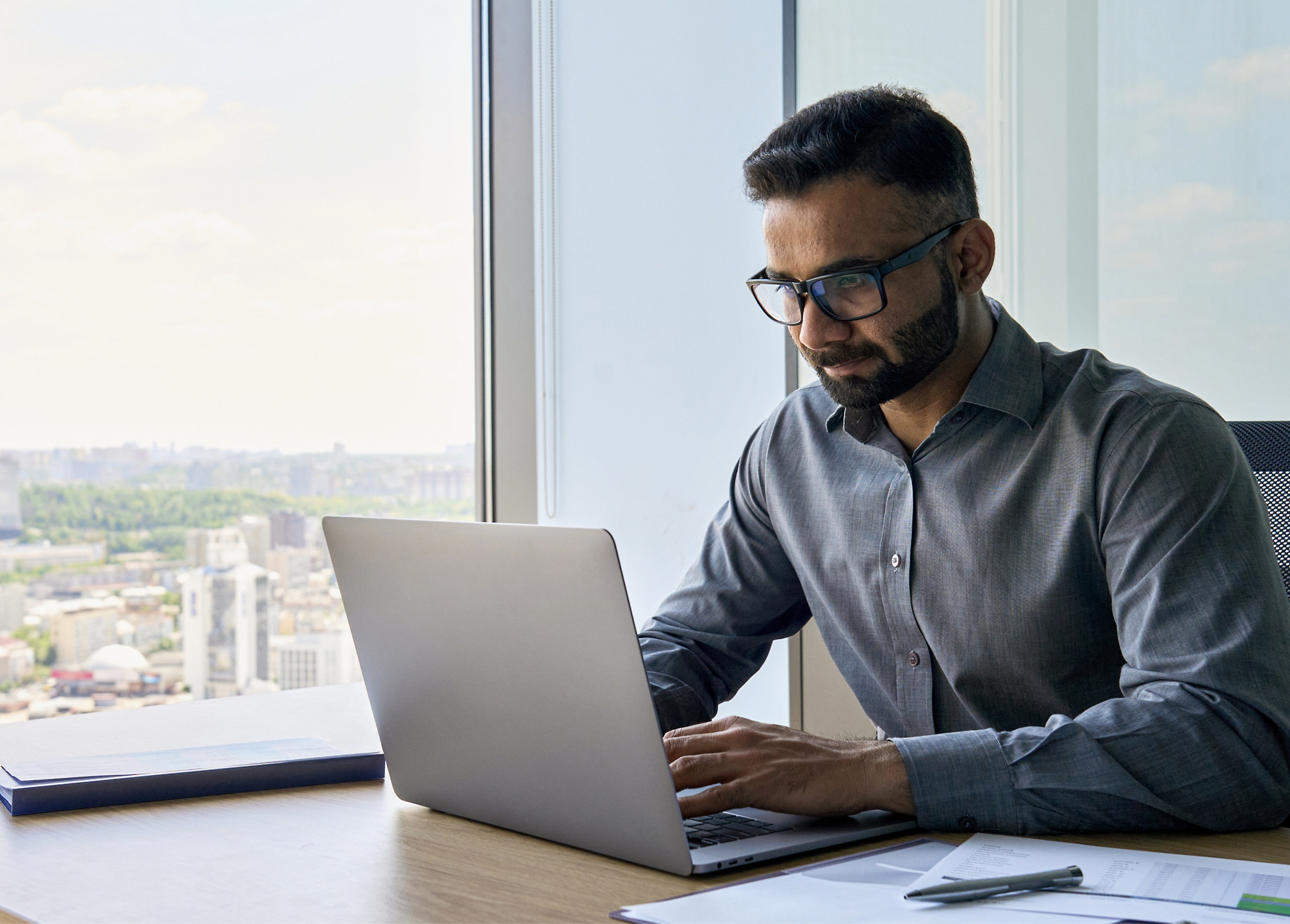 A man sitting at a desk by a large window, working on a laptop