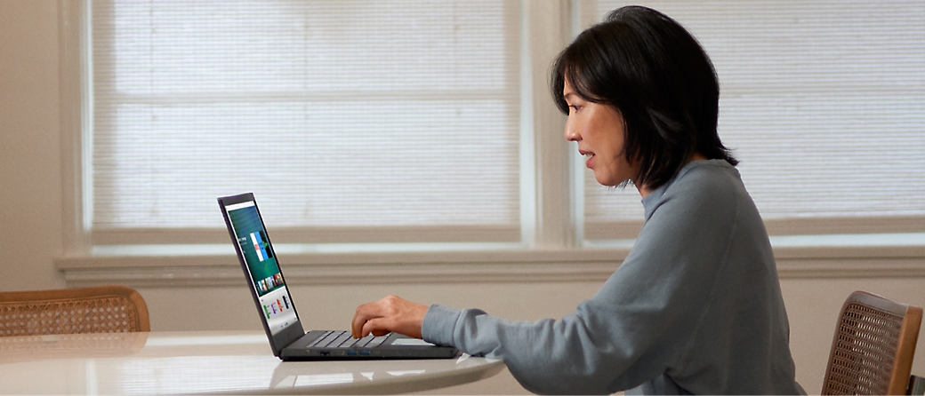 Eine Person, die einen blauen Pullover trägt und einen Laptop verwendet, während sie an einem runden Tisch in einem hellen Raum mit zwei Fenstern sitzt.