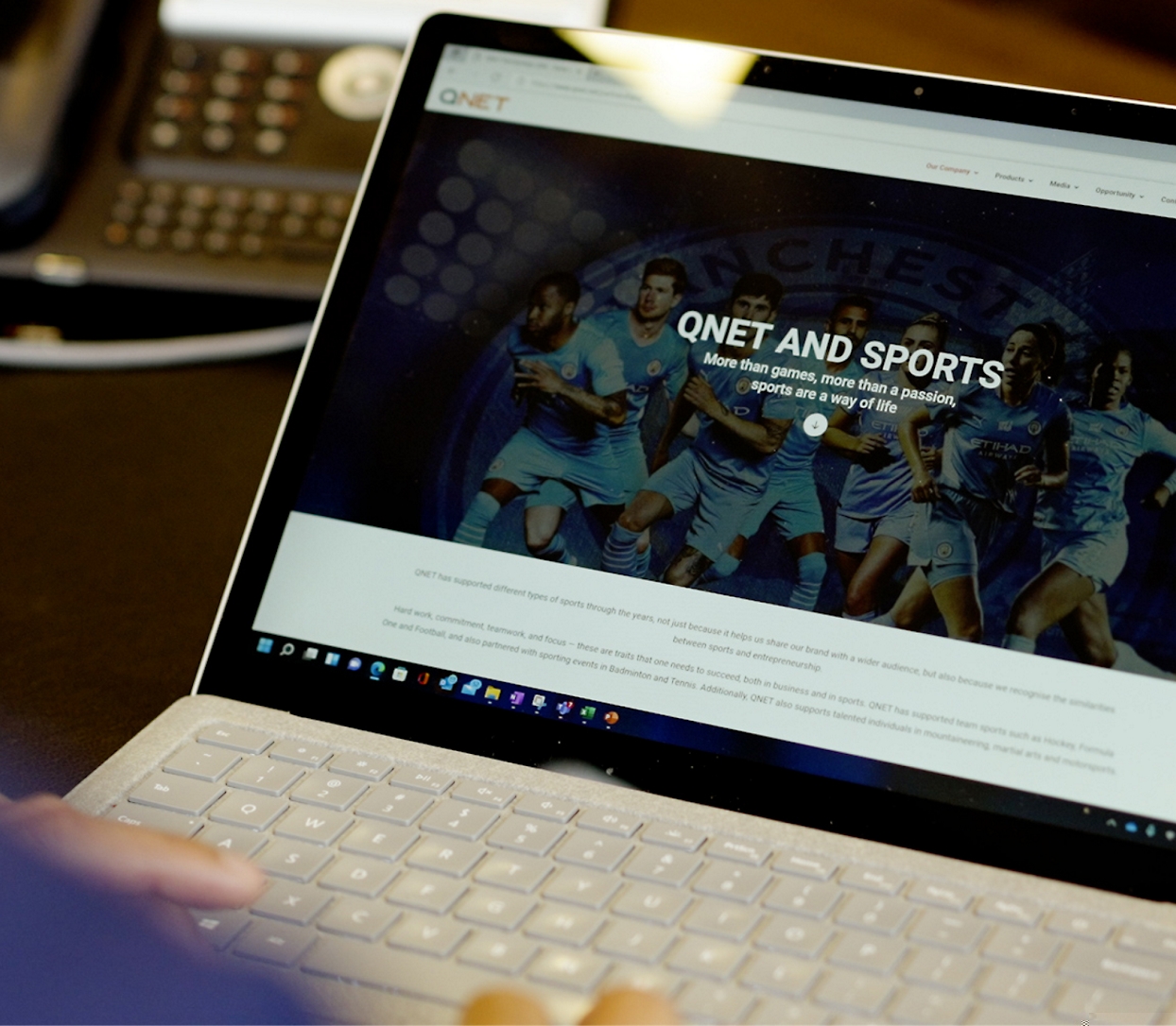 A person working on a laptop displaying a webpage titled "QNET AND SPORTS" with images of athletes and sports information.