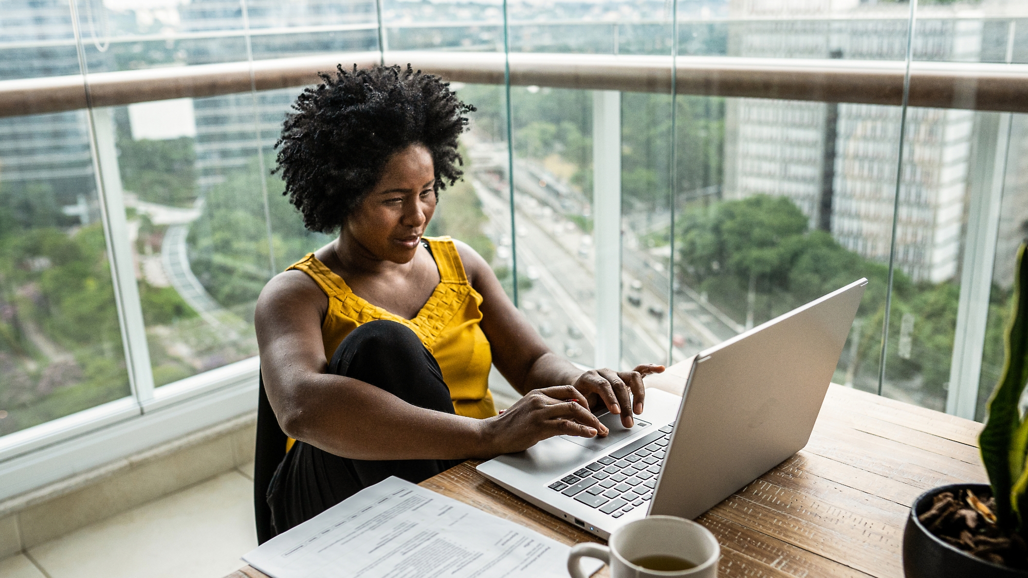 A woman works on a laptop at a desk by a window overlooking a cityscape, with documents and a coffee cup nearby.