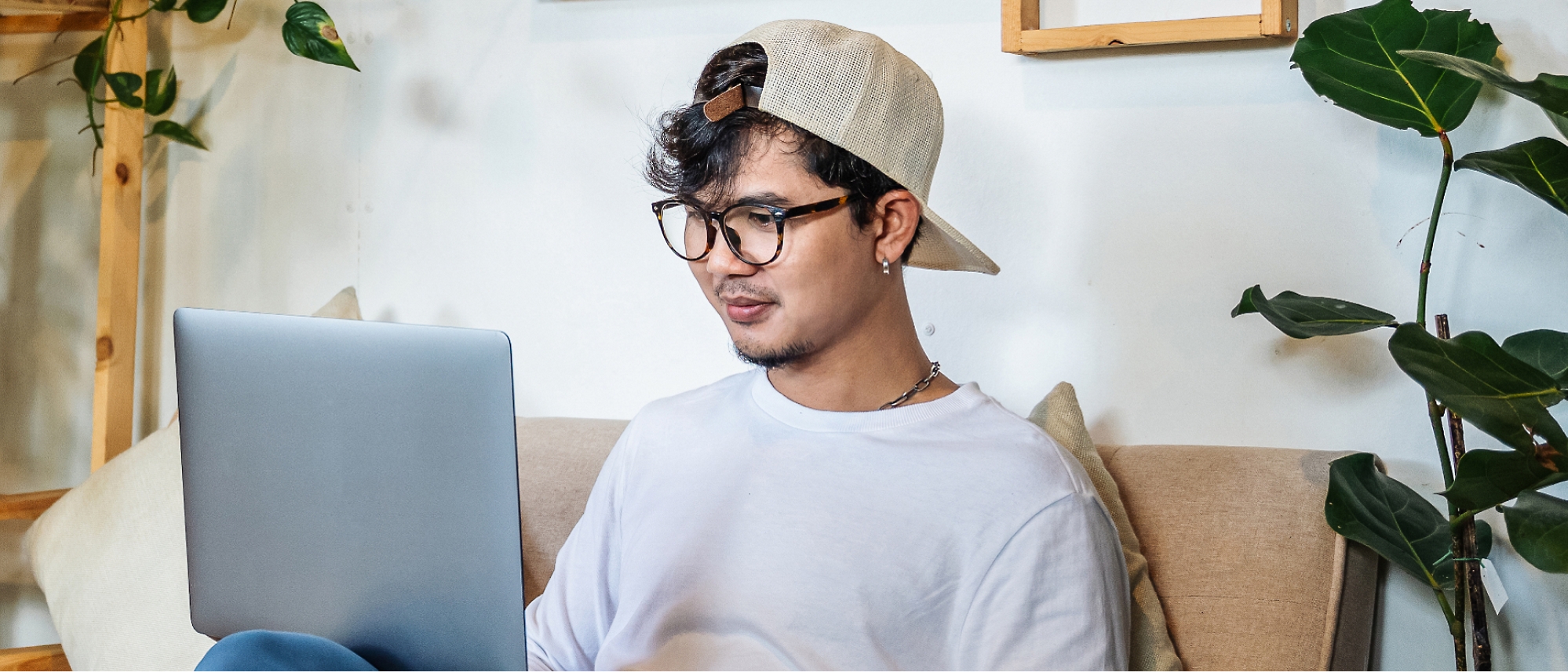 Uma pessoa usando óculos e um boné para trás está sentada em um sofá, usando um laptop