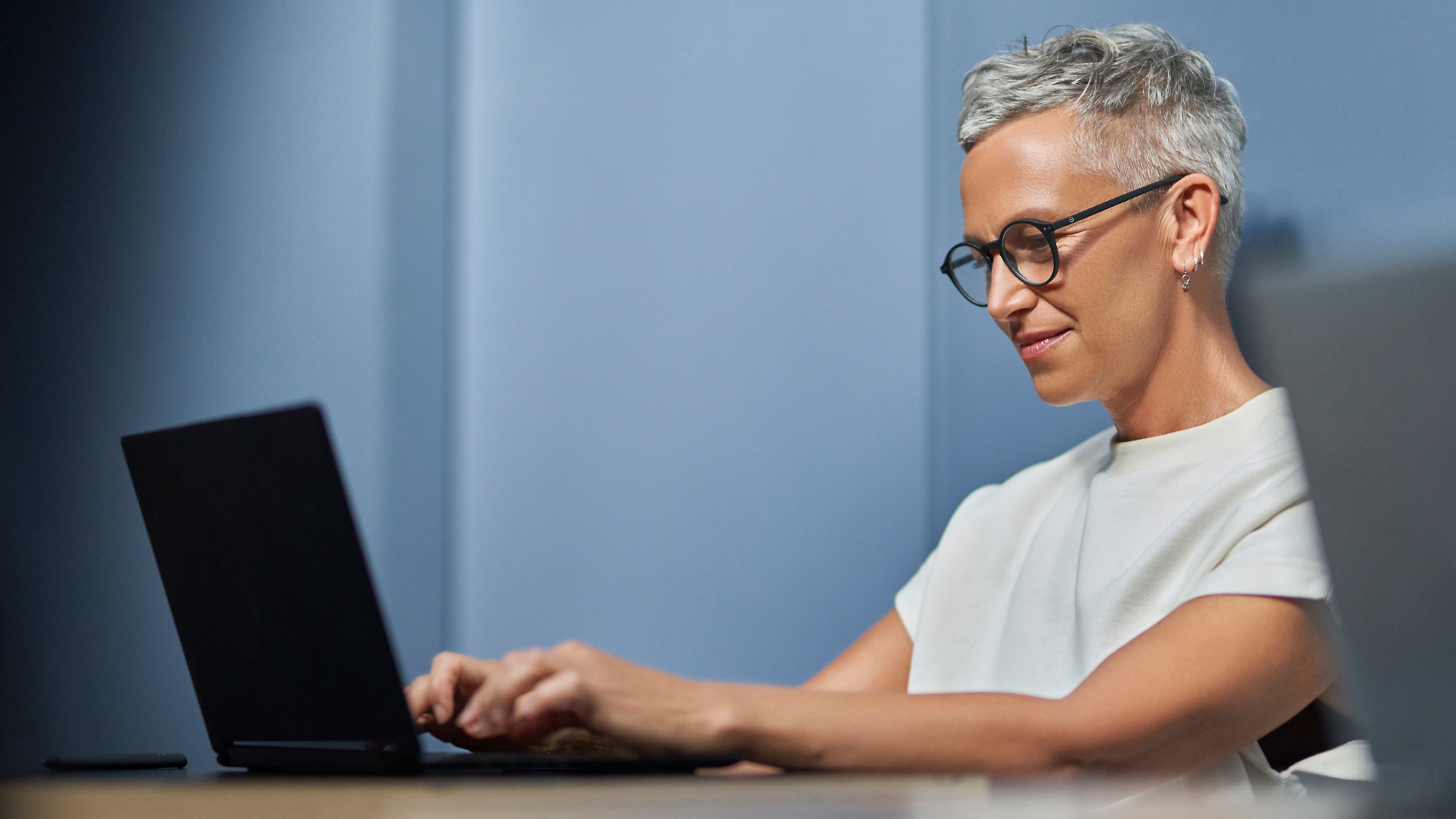 A mature woman with short gray hair smiling and working on a laptop in a modern office setting.