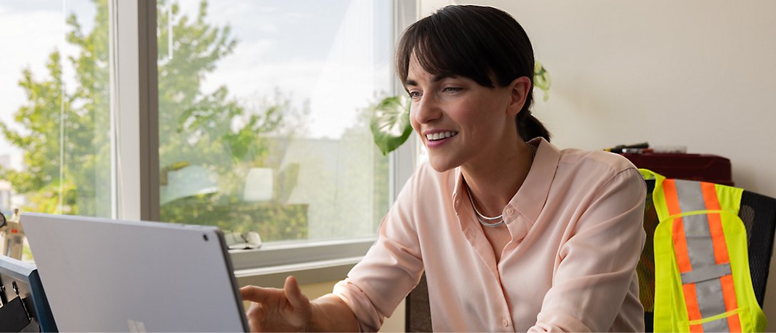 Woman in a pink blouse working on a laptop with a window and a safety vest in the background.