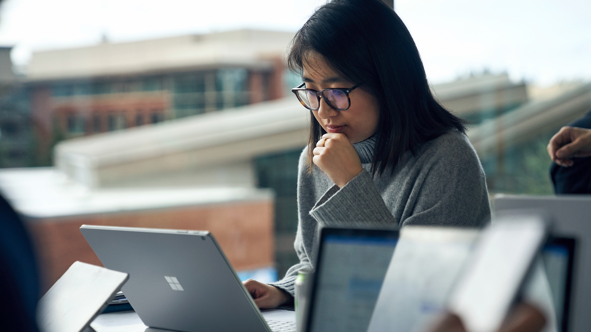 A focused young asian woman wearing glasses uses a laptop at a busy office table, with other workers